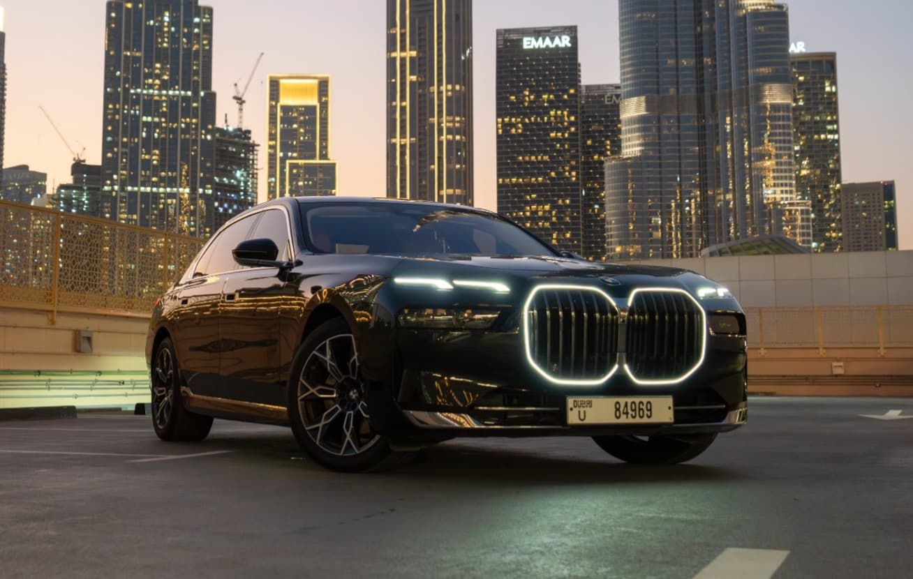 Вид спереди сбоку на BMW 7 Series с подсвеченной решёткой радиатора на фоне горизонта Дубая в сумерках
